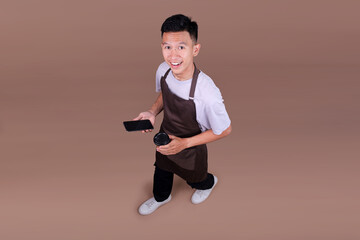Smiling Young Asian Man In Brown Apron Holding Smartphone And Coffee Cup