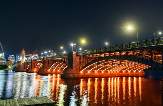 Night view of the Longfellow Bridge in Boston glowing with orange lighting, arching over the Charles River with reflections shimmering on the water and the city skyline in the background