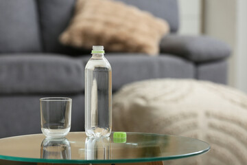 Bottle of fresh water and glass on table against stylish sofa in room