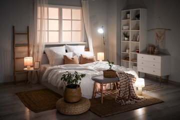 Interior of stylish bedroom with bed, shelving units, bench and glowing lamps at night
