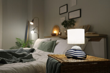 Glowing lamp with sleeping mask on wicker table near bed in bedroom at night