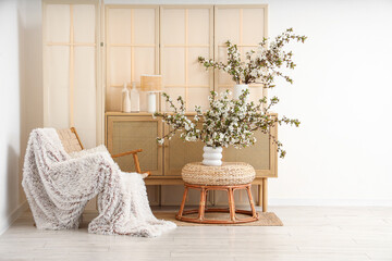 Interior of stylish living room with armchair, folding screen, commode and blooming branches in...