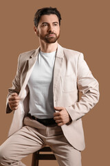 Handsome young man in light suit sitting on stool against beige background
