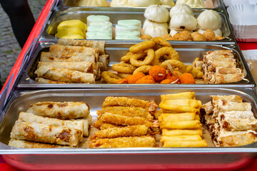 variety of Asian snacks displayed in metal trays at a festival food stall – including spring rolls, breaded shrimp, calamari rings, steamed baozi buns, sesame balls, and colorful rice rolls.