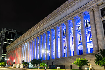Nighttime view of the New York State Education Building in Albany with its long Corinthian colonnade dramatically lit in blue and red. The Beaux-Arts facade stands against a dark sky, creating a