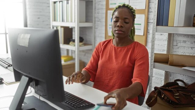 Woman working with computer indoors in office, wearing braids and red shirt, holding phone, showcasing focused and expressive demeanor.