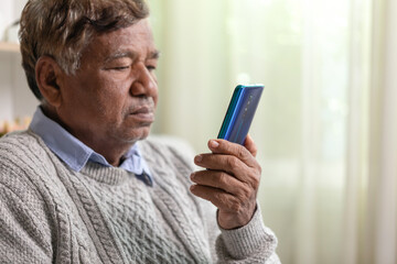 Sad elderly Asian man using mobile phone while sitting on sofa at home.