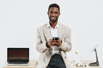 Professional young man using mobile phone in modern office environment, dressed smart casual, smiling confidently, with laptop and stationery on desk, white background, business and technology concept