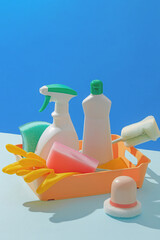 Bright and playful photo showing plastic cleaning bottles and tools inside a yellow basket on a blue backdrop, ideal for advertising cleaning products. Product bottle unlabeled, blank space for design