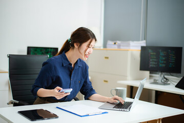 Young Asian female developer working on code with laptop and dual screens in a bright office. Ideal for themes of women in tech, programming