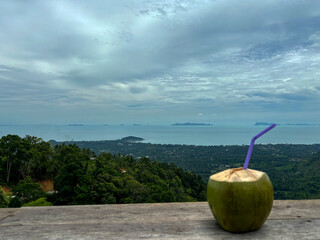 Coconut with a drinking straw on a wooden table with a view of Samui island from the terrace of the house on Lipa Noi Hill, Koh Samui