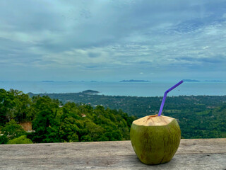 Coconut with a drinking straw on a wooden table with a view of Samui island from the terrace of the house on Lipa Noi Hill, Koh Samui