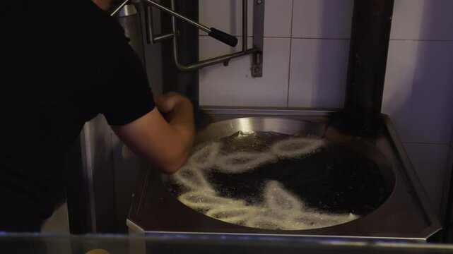 Guy places raw churros into boiling oil with hand. Dough sinks from special tool while oil bubbles and fryer fills with ring-shaped churros spirals