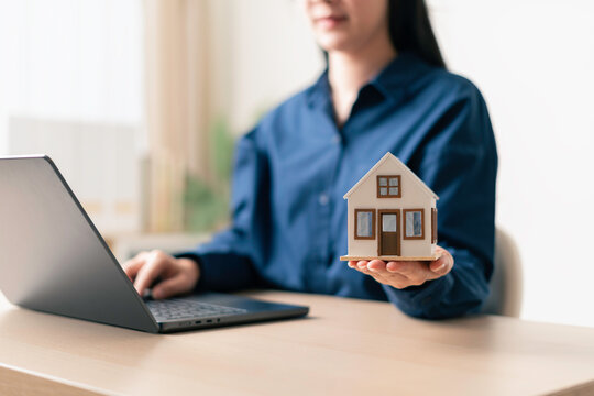 woman holding a house model while browsing property listings on a laptop in a home office setting for online real estate search