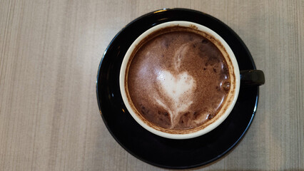 Top view of a cup of hot chocolate with heart-shaped latte art on a wooden table. Cozy and warm beverage in a black ceramic cup and saucer.