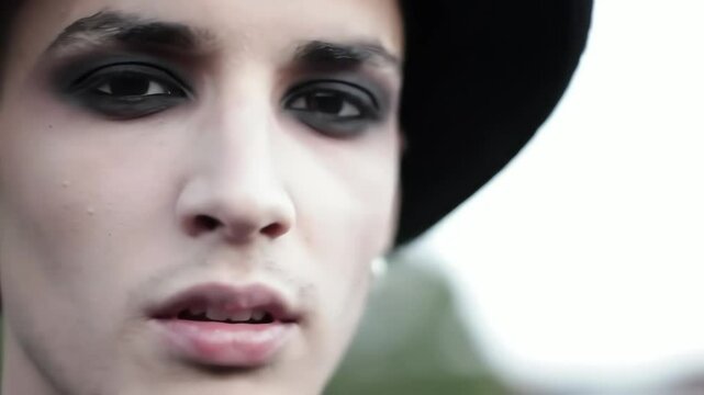 Young male model with dark smoky eye makeup and hat looking intensely outdoors close up shot