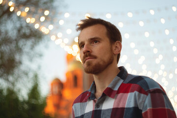 Thoughtful young man under festive outdoor city lights. Guy with beard and modern hairstyle wearing in blue, red and white plaid shirt walking through the city streets in urban background