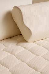 A realistic close-up photo of an orthopedic memory foam pillow resting on a quilted and textured mattress surface. The materials are light beige in color, with small perforated dots across the pillow 