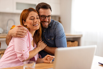 Obraz premium A happy couple working together on a laptop while reviewing documents. They share a moment of collaboration and planning, enjoying a productive and positive atmosphere in their well-lit living space.