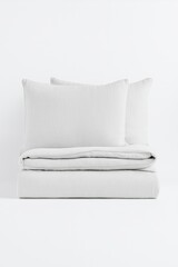 A perfectly folded white bedding set with two matching pillows, displayed in a minimalistic studio setting. The fabric is soft cotton with a light crinkled texture, conveying elegance and comfort. 