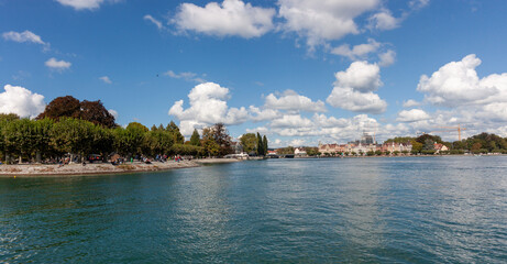 Fototapeta premium .A sweeping panoramic view of the serene Lake Constance (Bodensee) in Konstanz, Germany, under a brilliant blue sky dotted with scattered white clouds