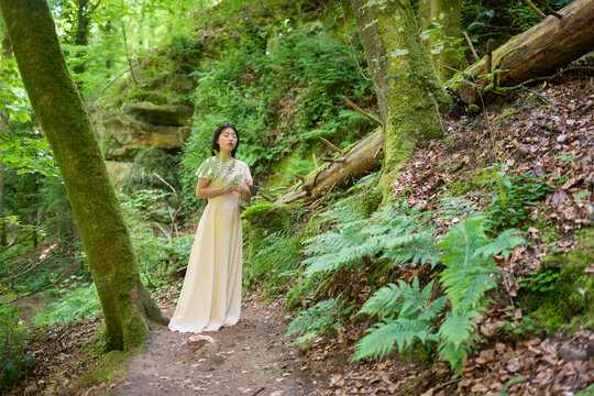 Naklejki art portrait of young asian woman in classic white dress a forest holding large fern leave on mullerthal trail in luxembourg