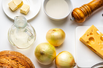 Ingredients for French onion soup. On the white table there is cheese, onions, bread, broth, mills with salt and pepper. Top view.
