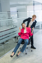 Coworkers fooling around in the office. A guy pushes a young woman around on a chair. 