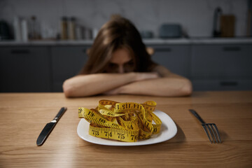 Depressed anorexic woman looking at plate with measuring tape served for dinner
