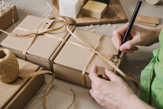 Person writes on a kraft tag attached to a ribbon-wrapped gift box near handmade soap. Handwritten tags, small business packaging, emotional branding, customer connection