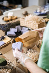 Person wearing gloves places lavender soap into a kraft bag among handmade skincare products. Handmade soap process, lavender skincare, indie soap maker, ethical manufacturing