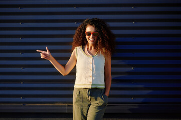 trendy woman wearing sunglasses pointing confidently against industrial metal wall in bright...