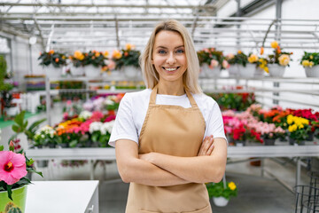 A smiling florist stands proudly with arms crossed in her shop, surrounded by vibrant blooms and greenery.