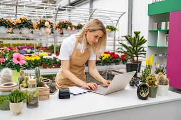 A Caucasian woman works on a laptop at a table in a flower shop surrounded by colorful plants and flowers.