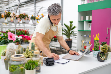 A Caucasian man in an apron works at the counter of a flower shop, using a laptop and taking notes while on the phone.