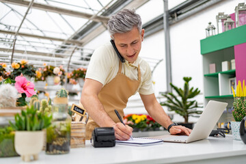 A Caucasian male shop owner takes an order over the phone while working on his computer at his...