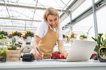 A Caucasian woman working in a florist shop, taking a call and using a laptop while writing.