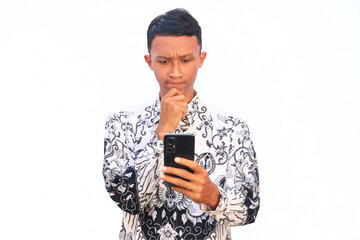 An Indonesian teacher in a PGRI uniform holds a smartphone and shows a thinking gesture.