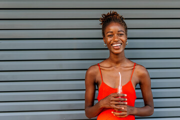 Young black woman enjoying a drink while standing against a gray wall during sunny afternoon