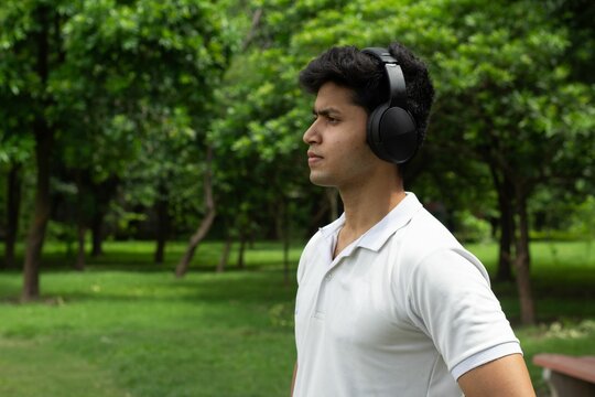Young man wearing headphones in a park, enjoying music amidst lush green trees.