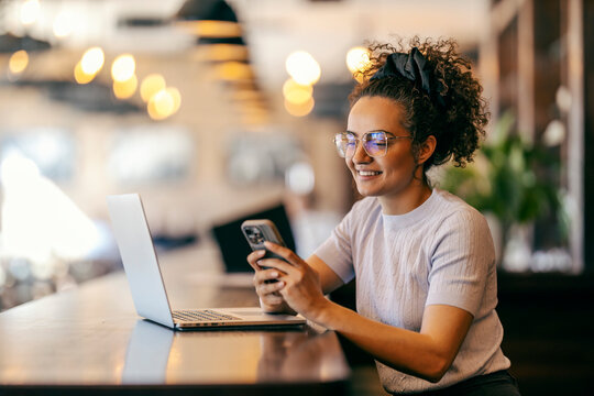 Cheerful interracial remote worker sitting in cafe with laptop in front of her and texting messages.