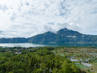 Aerial drone photo of Lake Batur and Mount Batur in Bali, with floating fish farms, green forest, and rural village. Serene natural landscape under dramatic cloudy sky