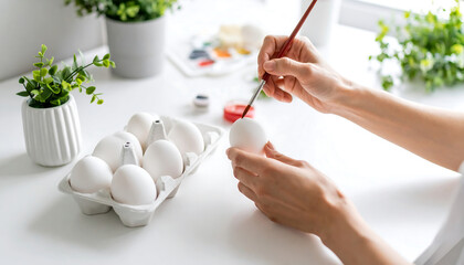 Hands painting a white Easter egg with a paintbrush, with a carton of eggs and art supplies nearby.