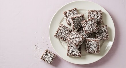 Top view of several lamington squares coated in chocolate and coconut, arranged neatly at the top center third. White plate with a pastel tablecloth background