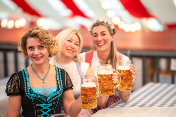 Women in traditional outfits cheer with large beer mugs at a wooden table in the beer tent. Oktoberfest Munich, Gäubodenvolksfest Straubing, Maidult Regensburg, Germany