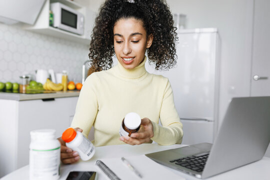Black girl restocking B-vitamins or poly-unsaturated fatty acids, making order online on marketplace holding bottles of biologically active supplements sitting at kitchen table in front of laptop