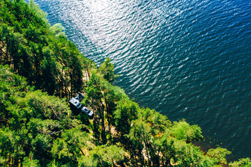 Aerial view of Caravan RV parked on the beach under a trees facing blue sea, lake or river. Travelers on an active family vacation.
