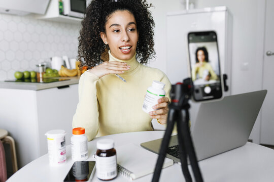 Black female blogger nutritionist recording video tutorial with advice and tips on immune boosting techniques on smartphone camera recommending food supplements, holding bottle in hand