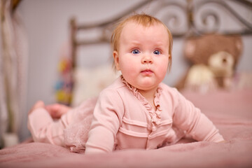 Fototapeta premium Baby girl with light hair and blue eyes lying on pink bed. Soft lighting and bedroom setting create warm and peaceful atmosphere