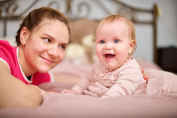 Young mother with brown hair, smiling baby girl indoors. Warm pink tones, soft lighting, metal bedframe in background. Expression of joy and connection